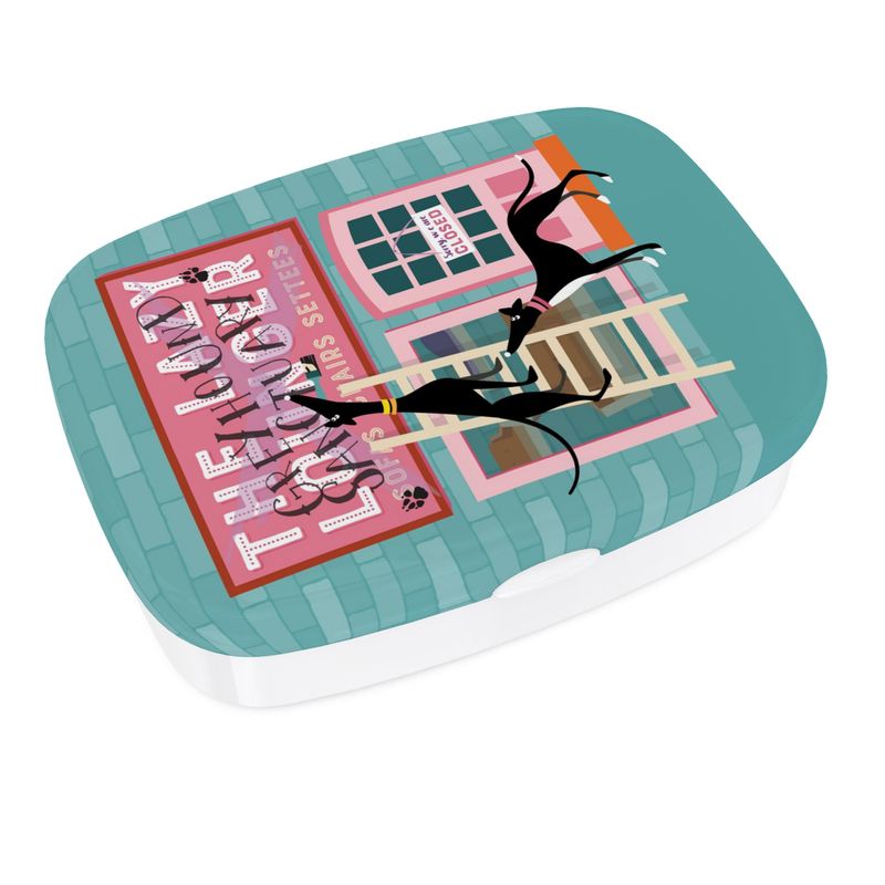 Different angled view of the lunch box. The base is white. The top has a full colour print wrapped around the sides. The background colour on the lid is turquoise. The image on the lid shows the front of a sofa shop called the Lazy Lounger. It is currently closed. There is a greyhound standing up a ladder with a paintbrush in their mouth. They have painted 'Greyhound Sanctury' over the top of the shop sign. There is a 2nd greyhound keeping watch.